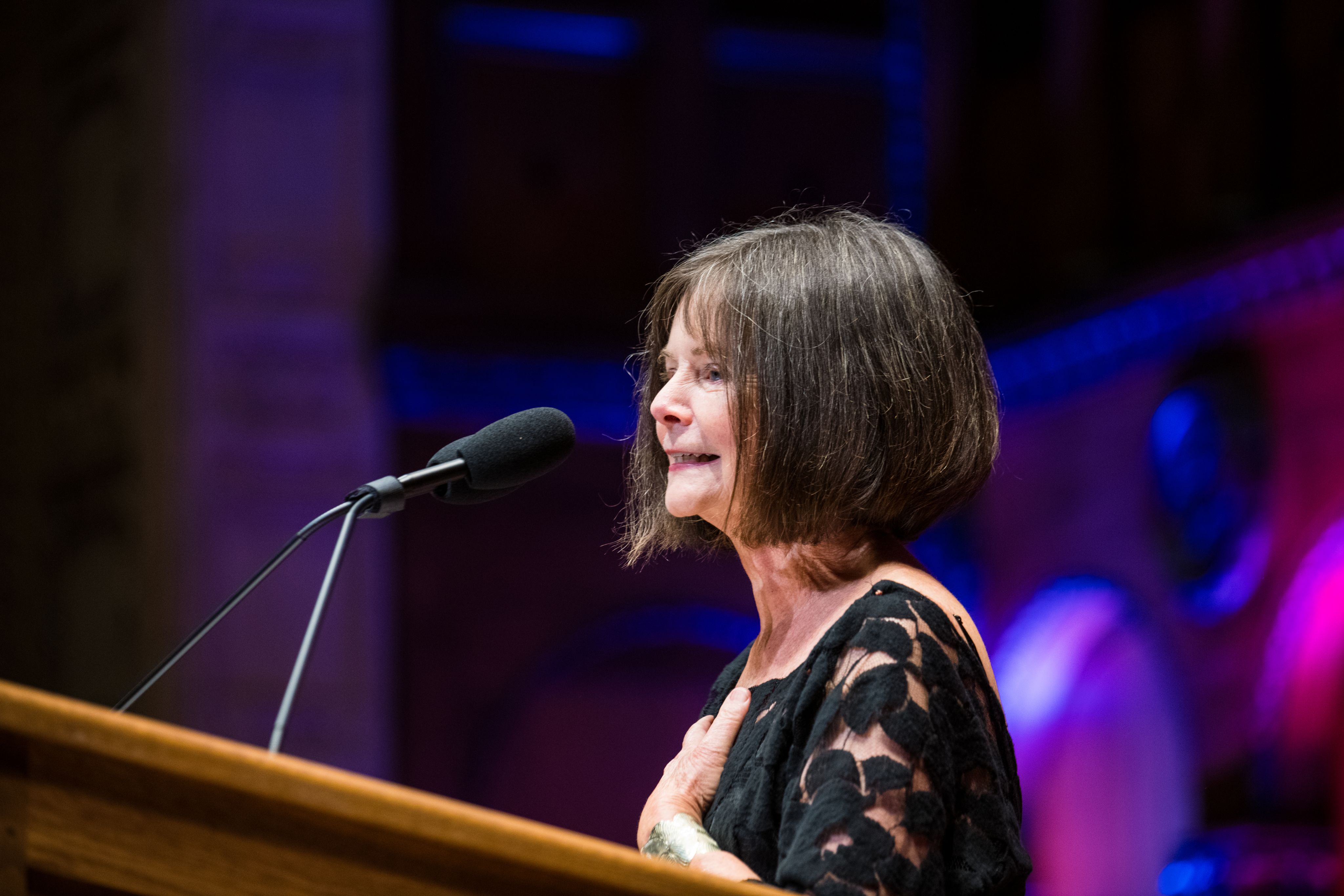 A woman with her left hand on her chest speaks into a microphone at a podium.