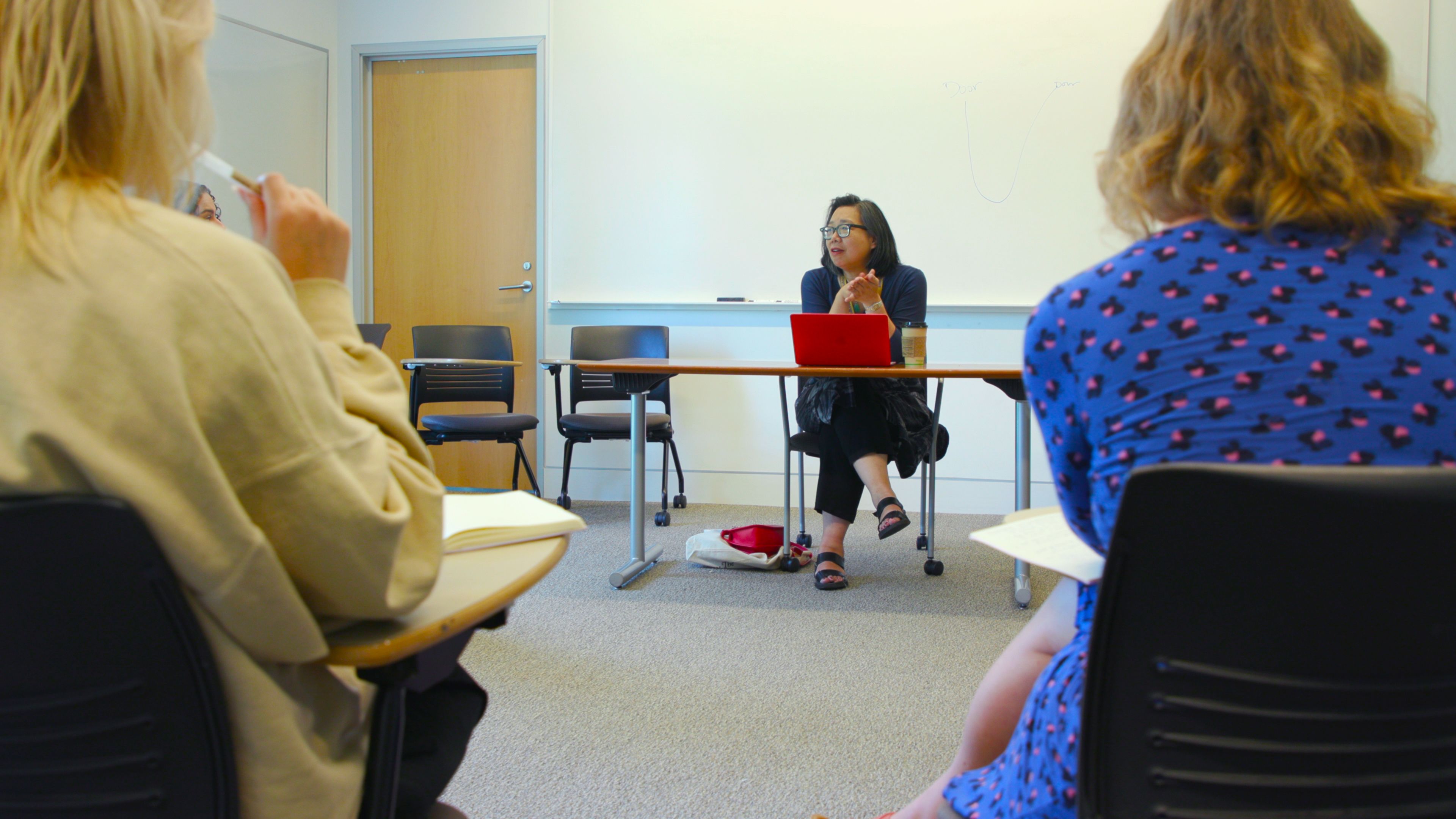 An Asian woman sits at a table behind a red laptop speaking to people sitting at desks in a circle.