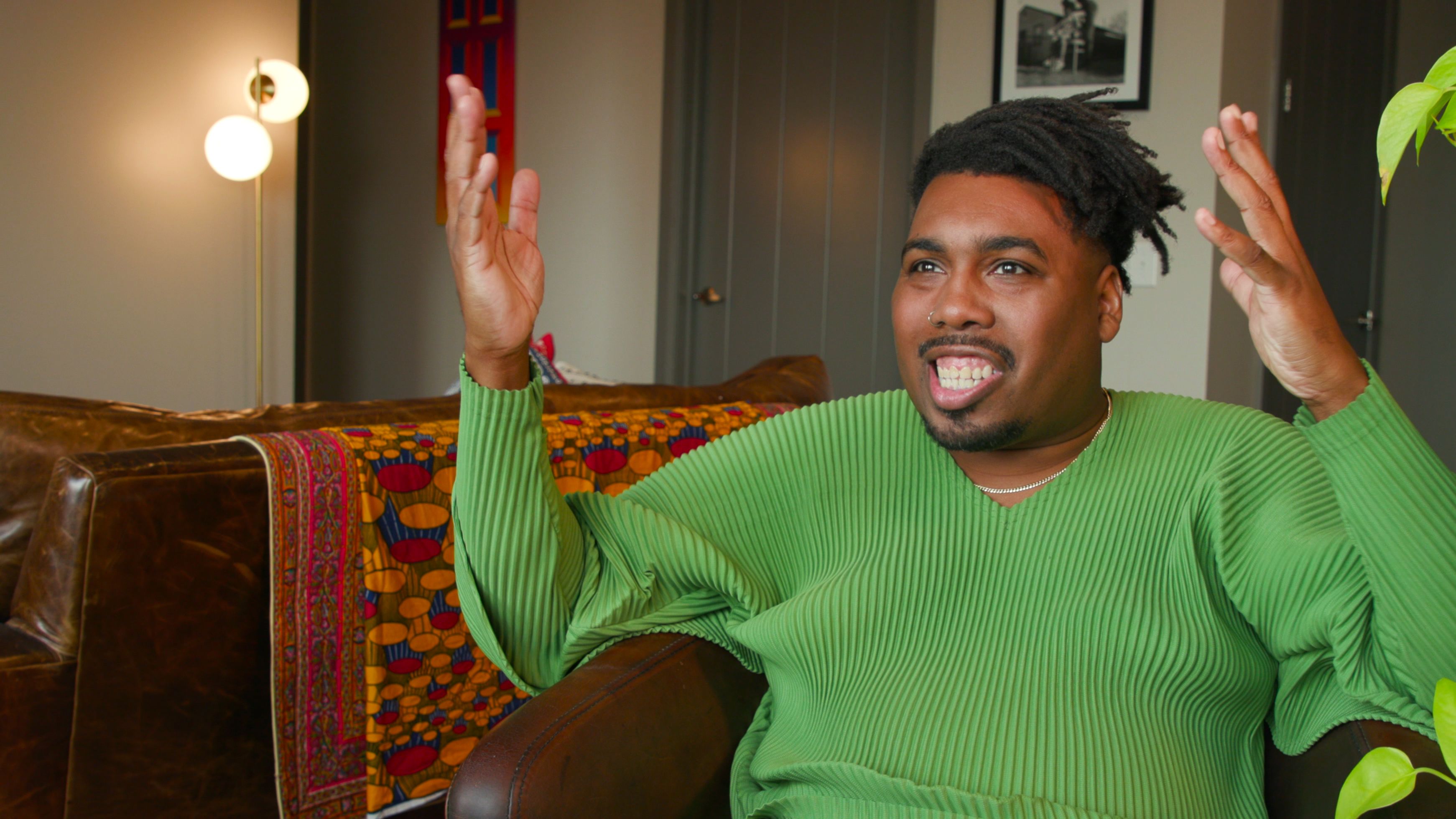 A Black man in a green sweater raises his arms to gesticulate as he talks while sitting.