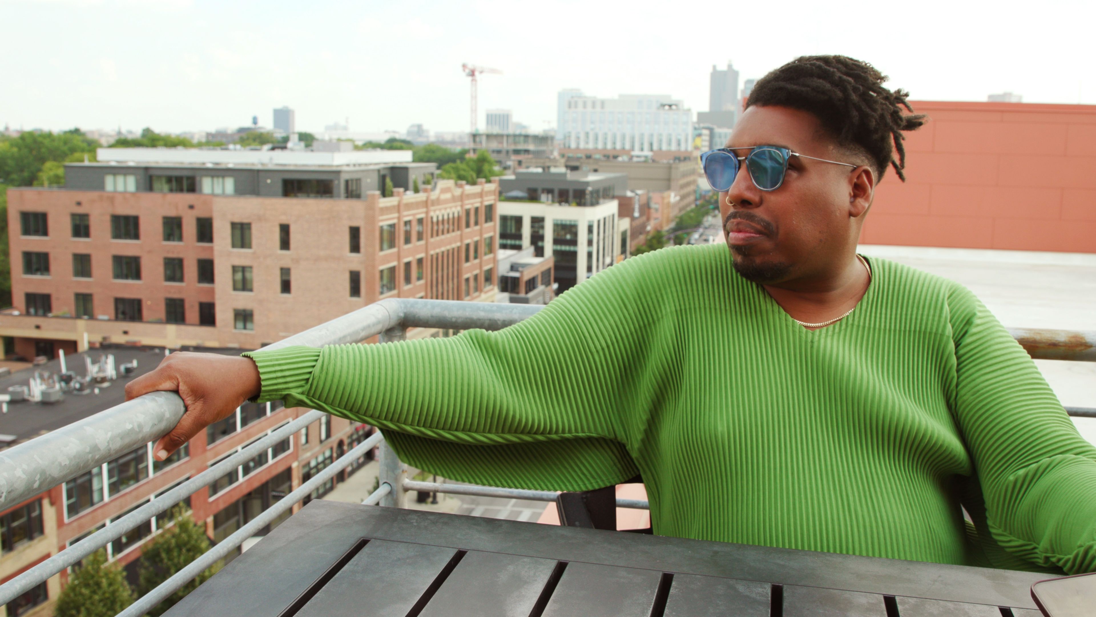 A Black man in sunglasses and green sweater sits at a table on a rooftop.