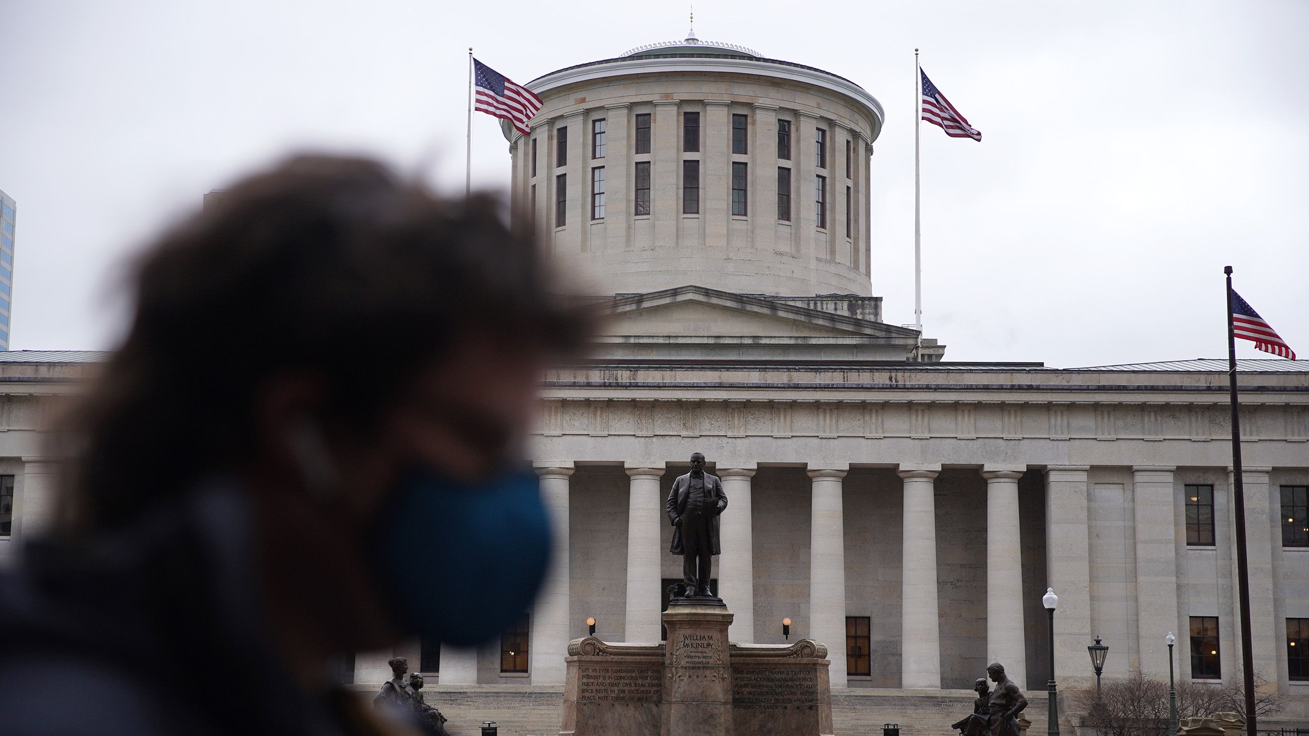 A person wearing a mask in the foreground walks past the Ohio Statehouse in Columbus.